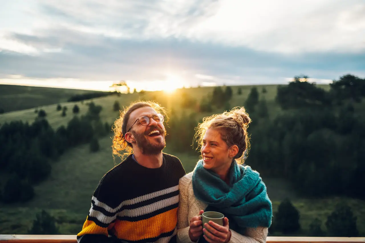 A couple laughing over a hot drink on a green hillside at sunset