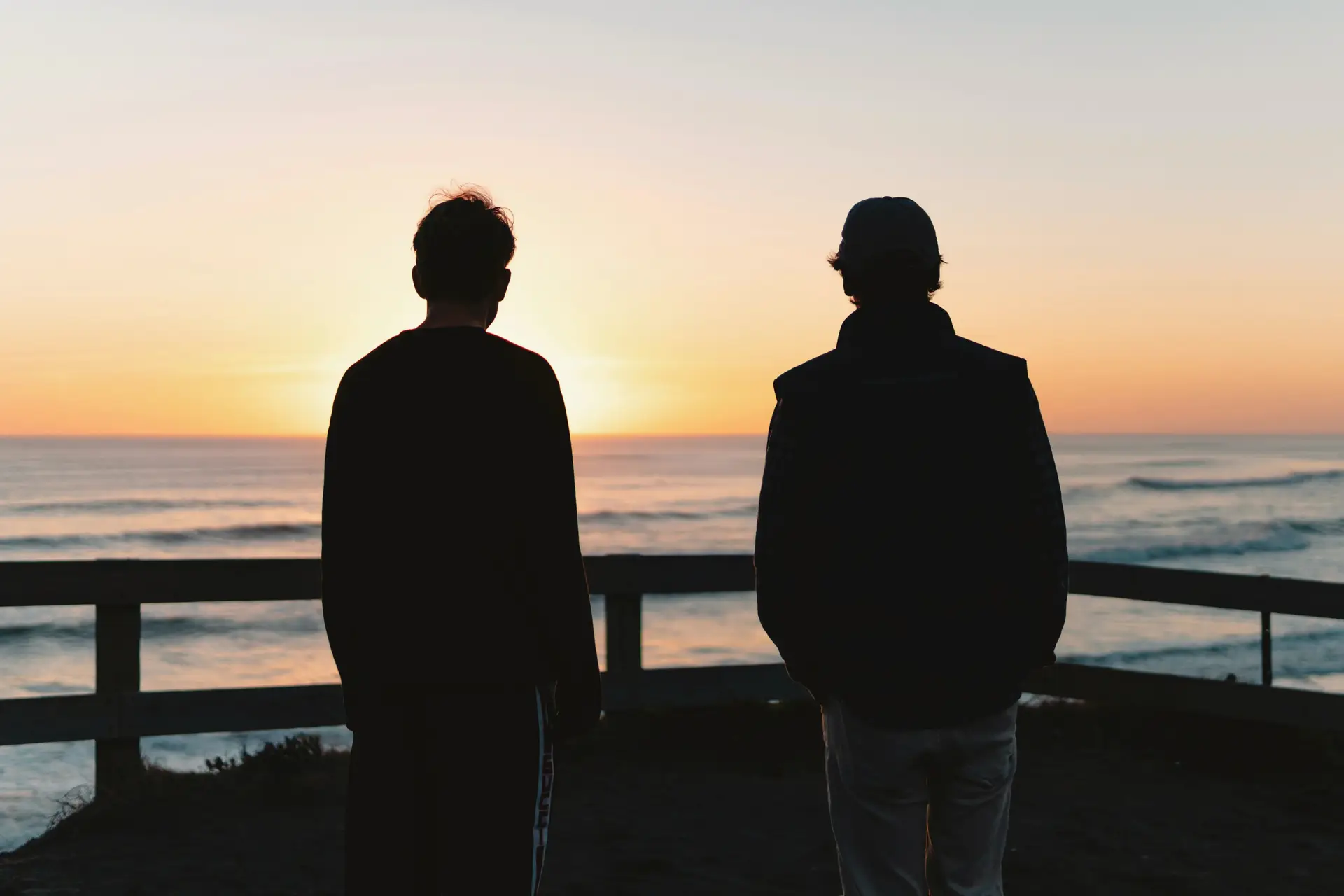 Two men at a beach lookout rail, watching the sun set over the ocean